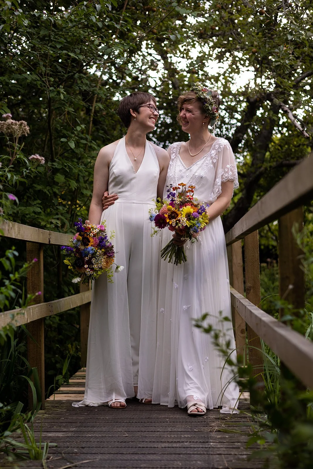 Two women in white dresses standing on a wooden bridge surrounded by greenery, smiling at each other, holding colorful bouquets, one with a floral crown.