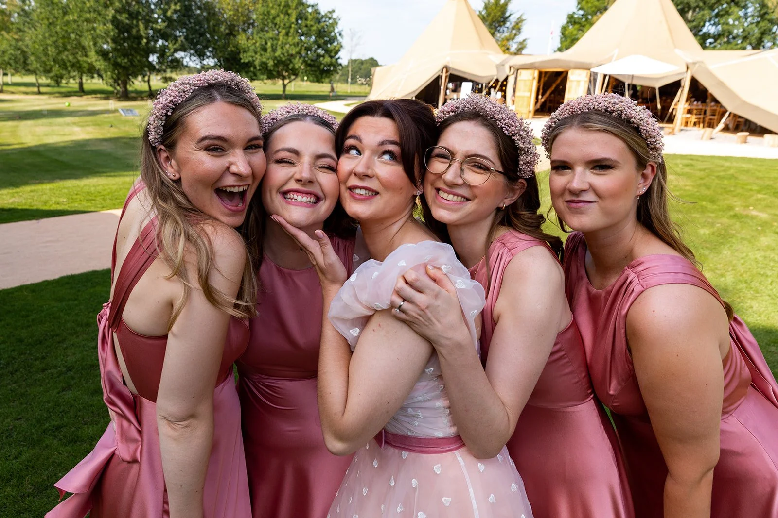 Group of five women celebrating a wedding outdoors, wearing pink dresses and floral headpieces, with a large tent in the background.
