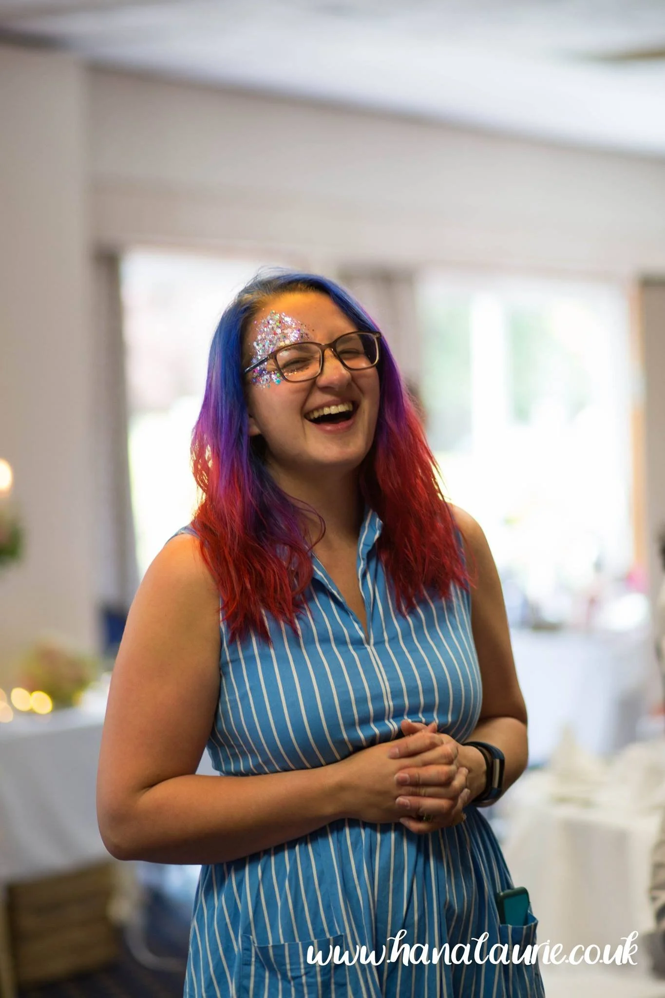 A woman with colorful hair, wearing glasses and a blue striped dress, smiling and laughing indoors.
