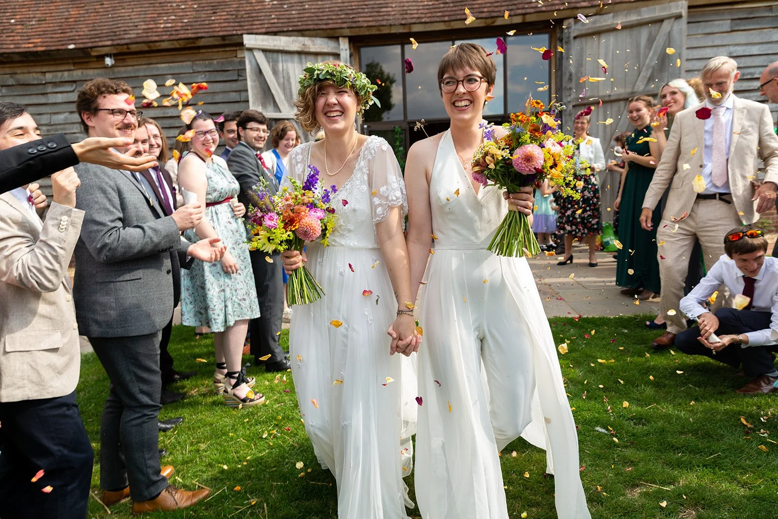 Two brides in white dresses holding hands and bouquets, surrounded by wedding guests throwing flower petals outside a rustic building with wooden siding.