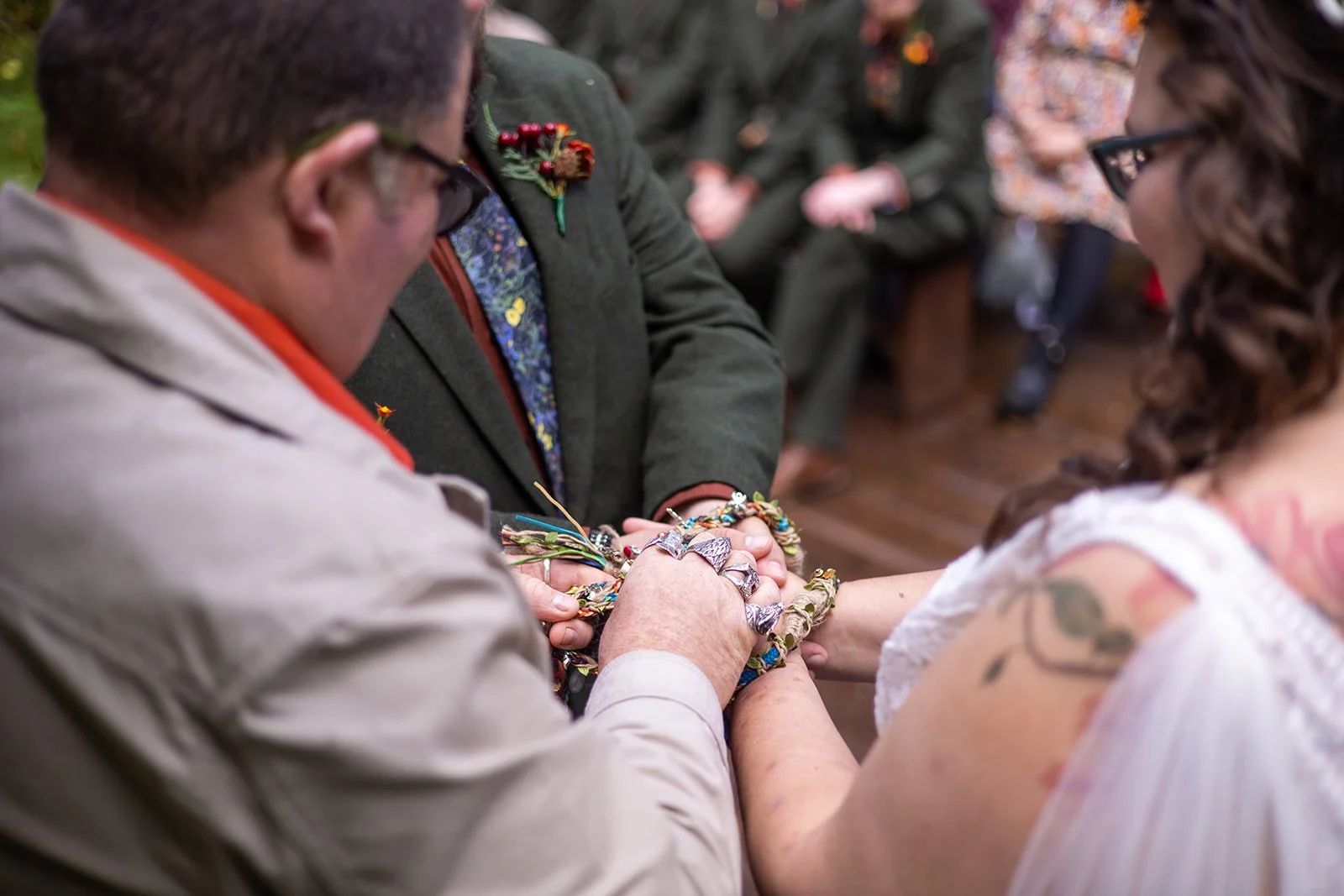 A couple and a officiant hold hands during a wedding ceremony, with the focus on their hands and rings, surrounded by jewelry and greenery.