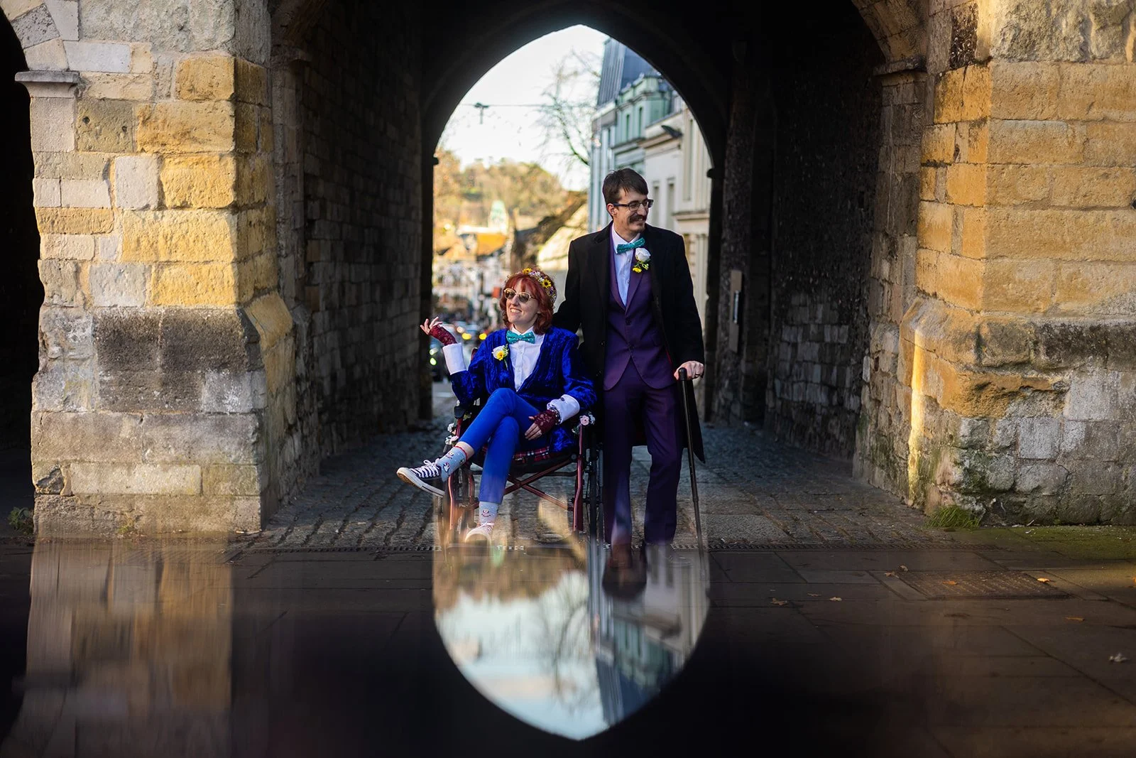 A couple with disabilities walks under a stone archway. The woman, sitting in a wheelchair, wears a blue velvet blazer and has red hair. The man, standing and pushing her wheelchair, wears a purple suit with a blue bow tie and has dark hair and glass