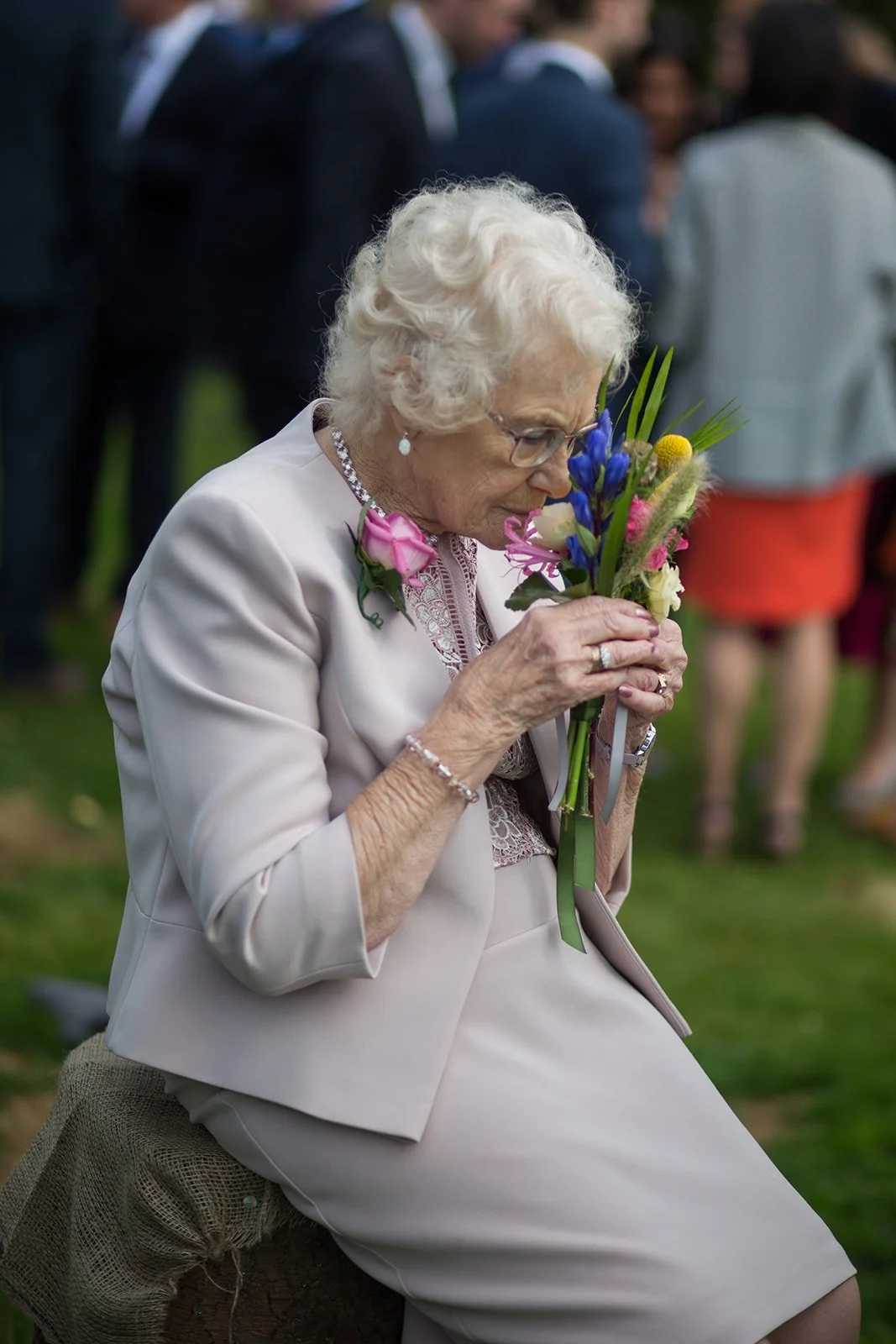 An elderly woman with white curly hair, glasses, and jewelry, sitting outdoors holding a bouquet of flowers close to her face, with a blurred crowd in the background.