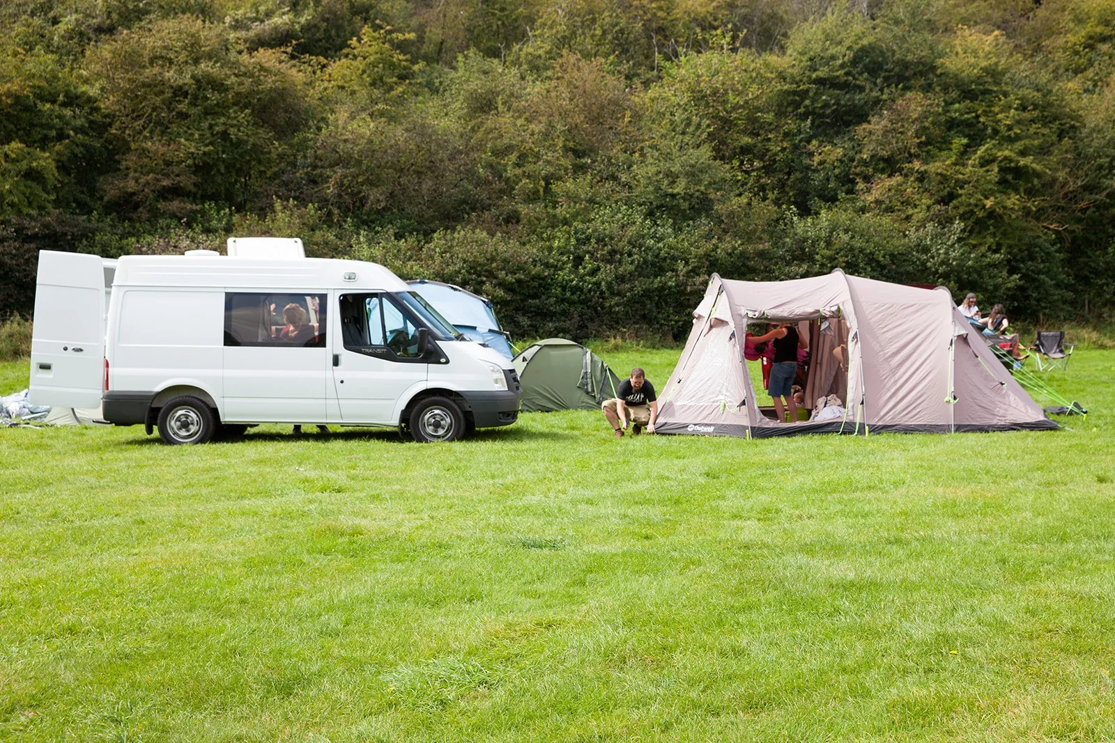 A group of people camping on a grassy field with a white van, tents, and outdoor chairs, surrounded by trees.