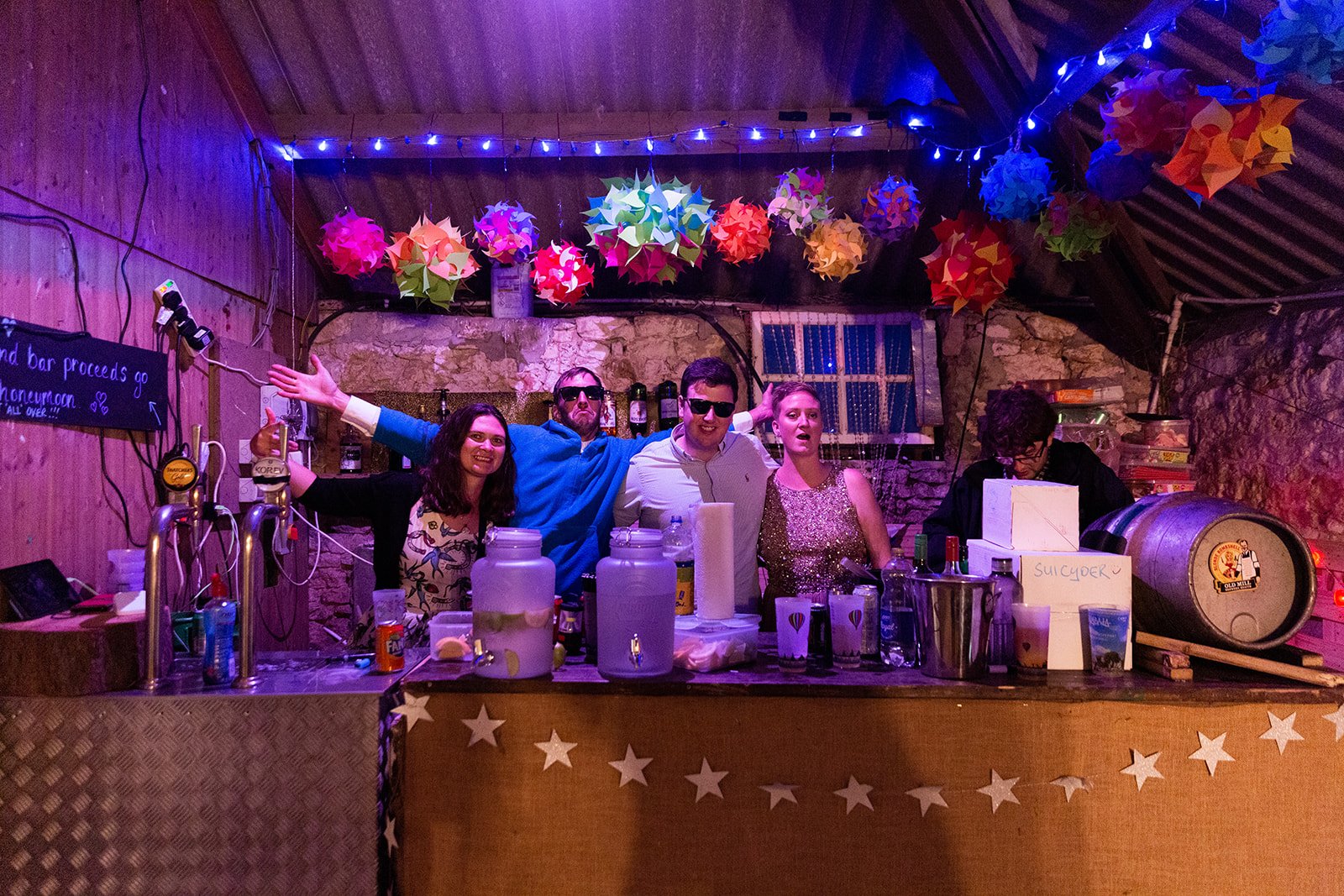 Five people standing behind a bar decorated with stars and hanging paper lanterns, with drinks and bar equipment on the counter, inside a rustic venue with purple and blue lighting.