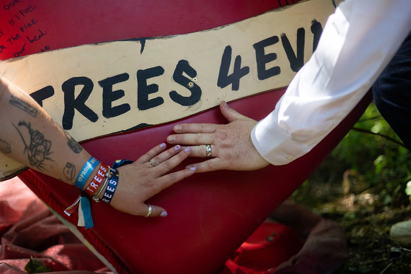 Two people with hands on a red box with a sign that reads "TREES 4 EV". One person is wearing bracelets with the word "TREES" and has tattooed arm. The other person is wearing a white shirt.