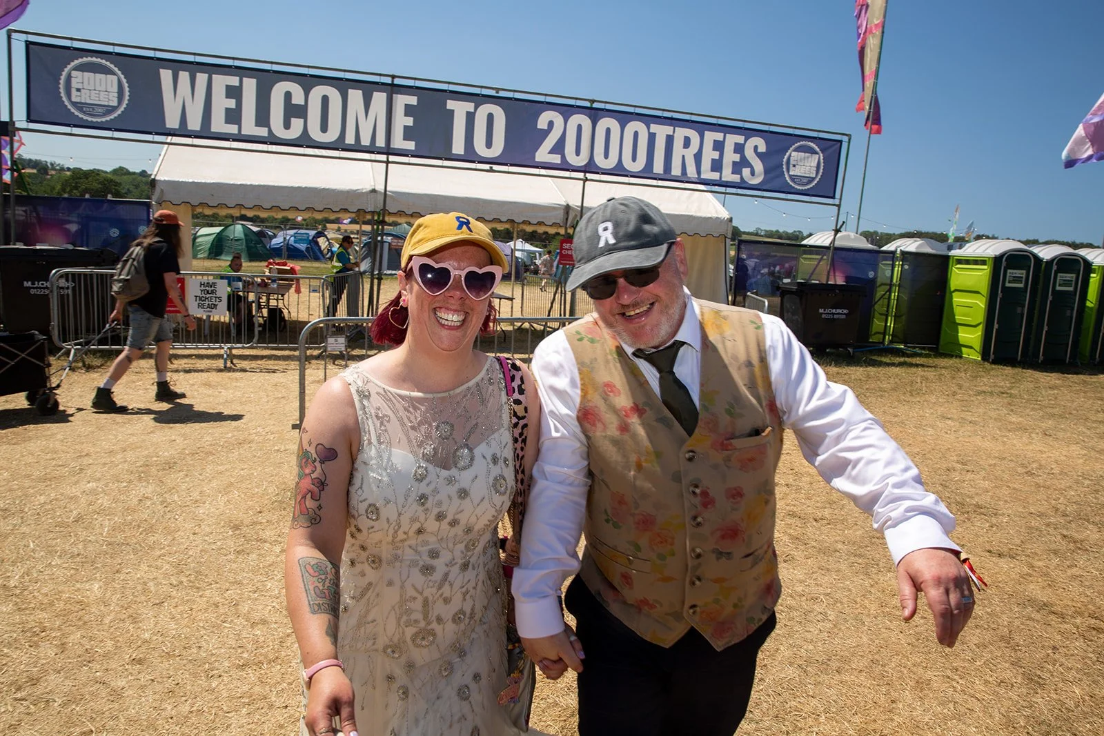Smiling woman and man holding hands at an outdoor festival with a welcome sign that says 'WELCOME TO 2000 TREES' in the background.