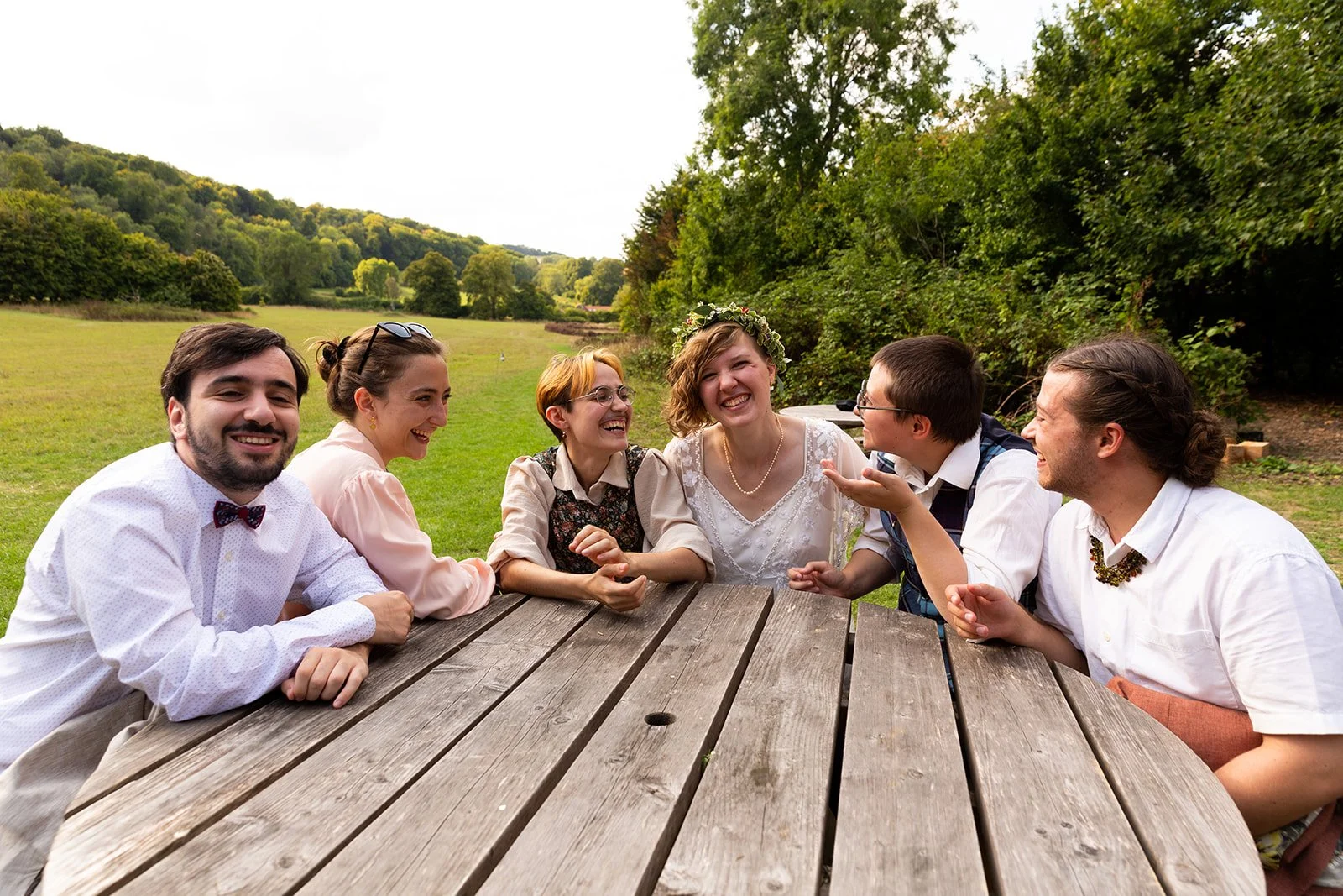 Group of seven friends sitting at a picnic table outdoors, smiling, and laughing together on a sunny day with trees and hills in the background.