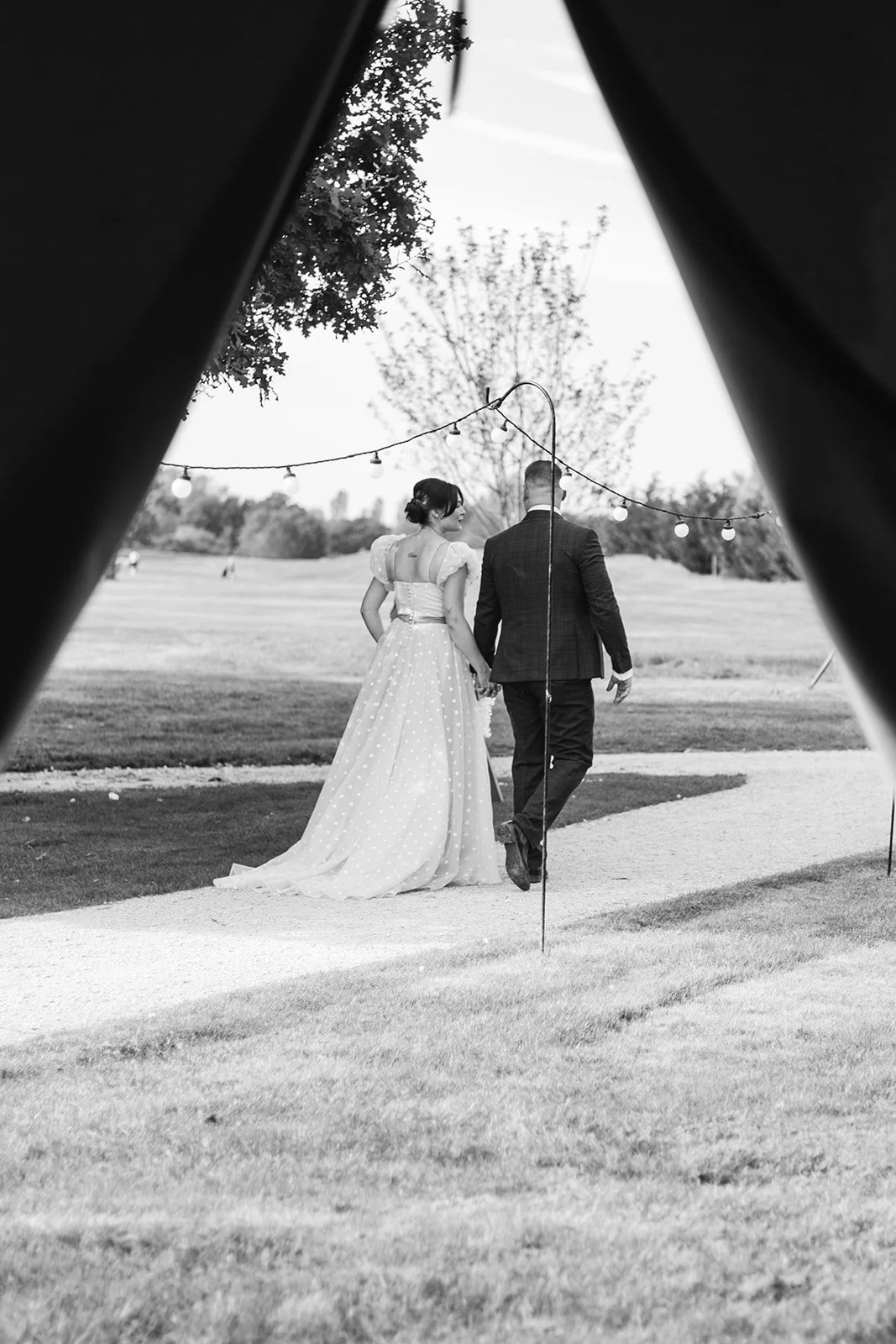 A black-and-white photo of a newlywed couple walking hand in hand outdoors, framed by a tent or canopy opening with string lights above, in a park with trees and open space.