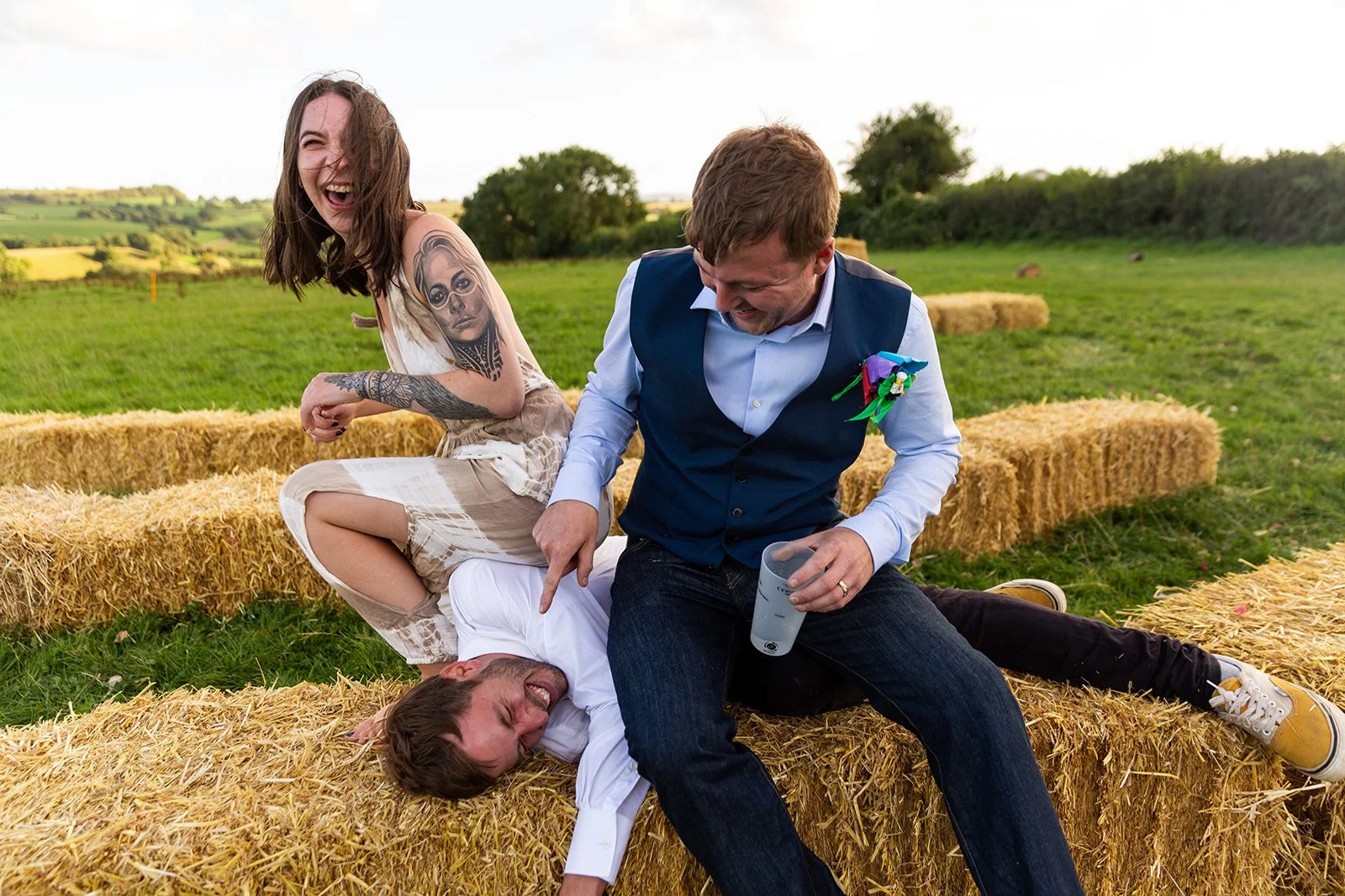 Group of four people having fun outdoors on hay bales, laughing and joking, in a rural field with trees and cloudy sky in the background.