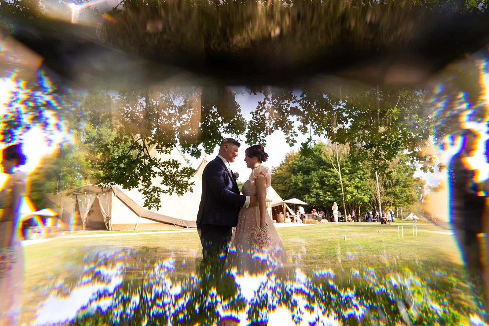 Couple at a wedding, standing under a tree, with a tent and outdoor reception area in the background, seen through a water droplet or kaleidoscope effect.