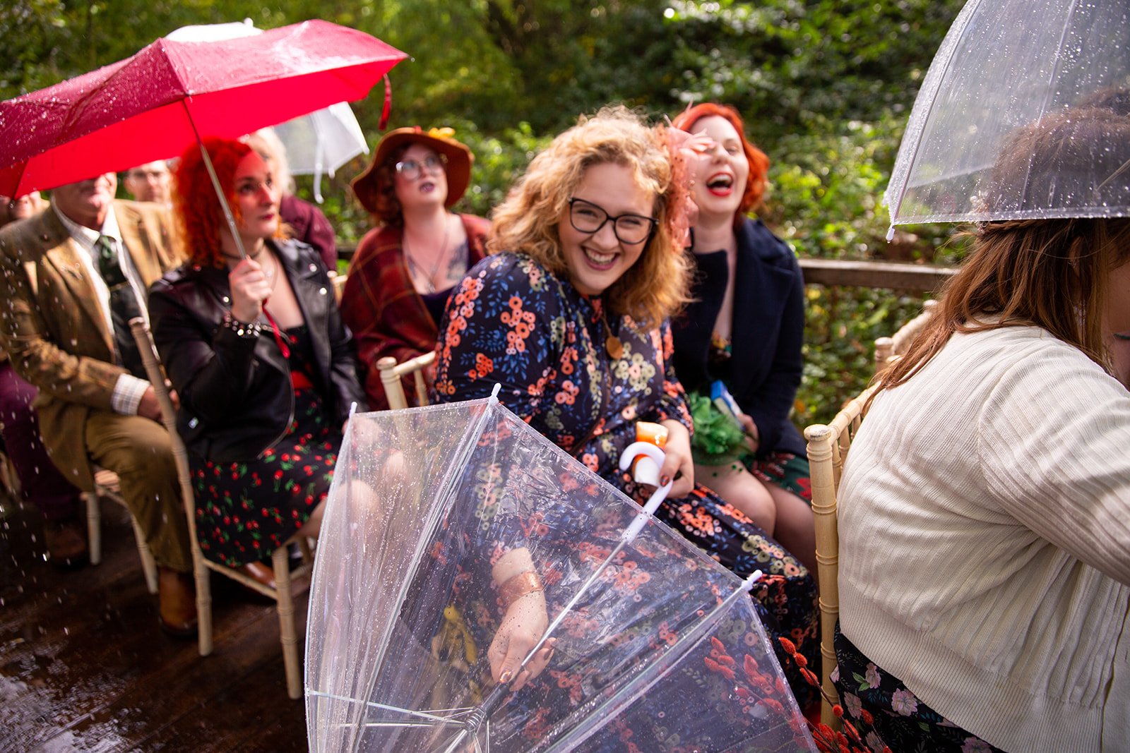 A group of people sitting outdoors under umbrellas, smiling and enjoying a rainy day, with some holding umbrellas and wearing colorful clothing and accessories.