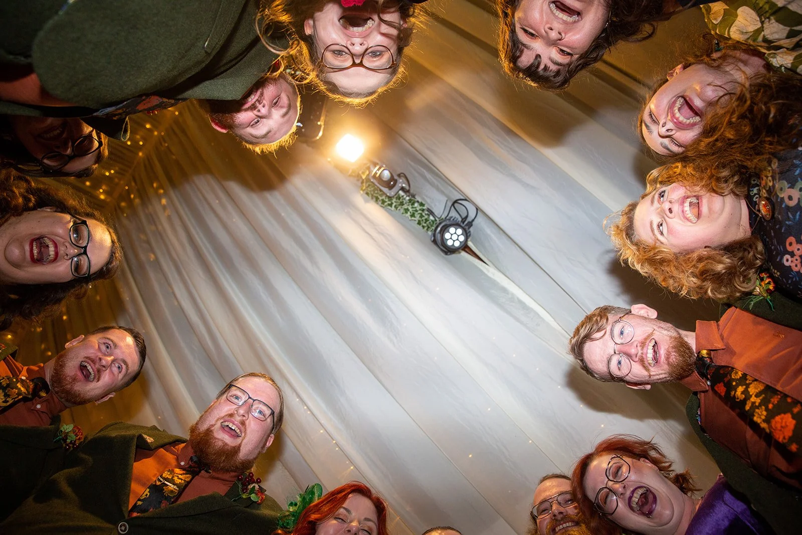A group of people gathered in a circle, looking down and smiling, with a white curtain background and a mounted light on the ceiling.