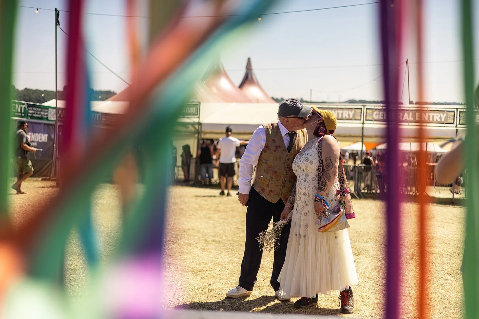 A couple sharing a kiss at a festival, seen through colorful carnival rides with tents and people in the background