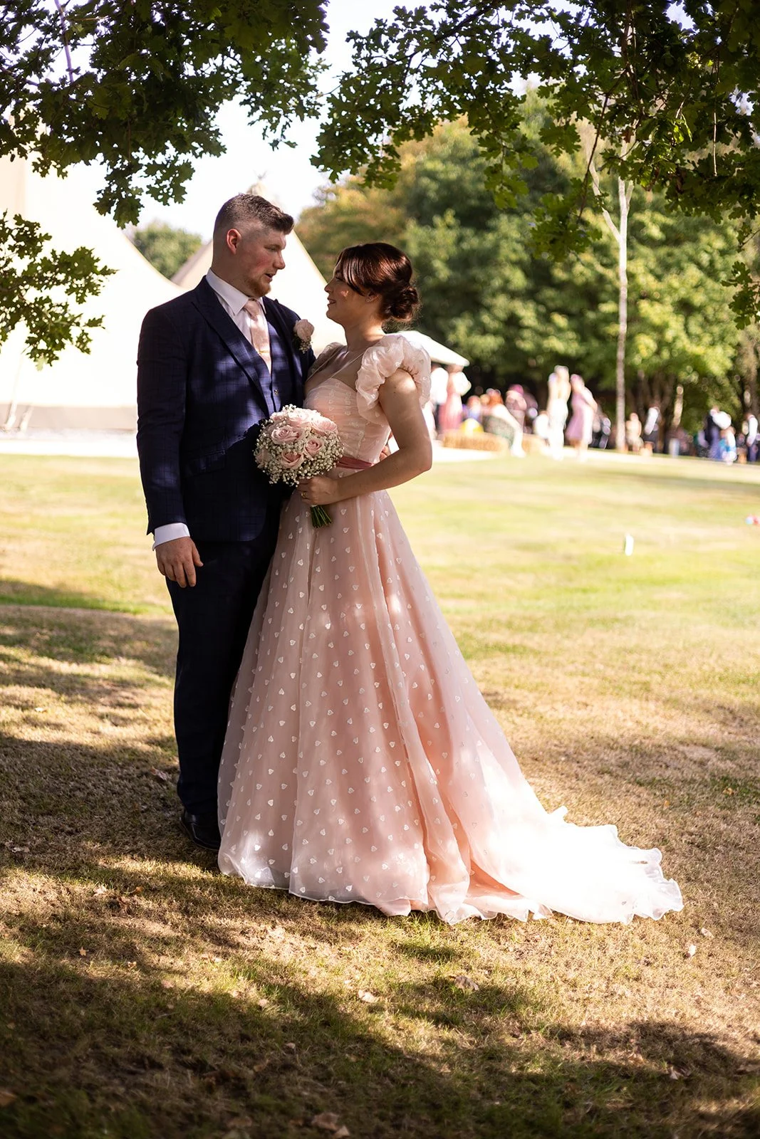 a couple standing embracing in front of a tipi at mad swans hampshire, they're in the shade of a tree and the bride is in a pink polkadot dress
