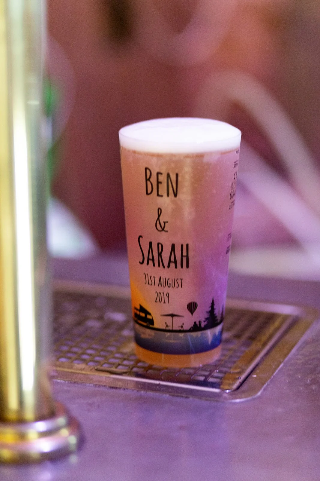 A personalized drink cup with the names Ben & Sarah, wedding date August 31, 2019, featuring a silhouette of a picnic table, a lake, trees, and a hot air balloon.