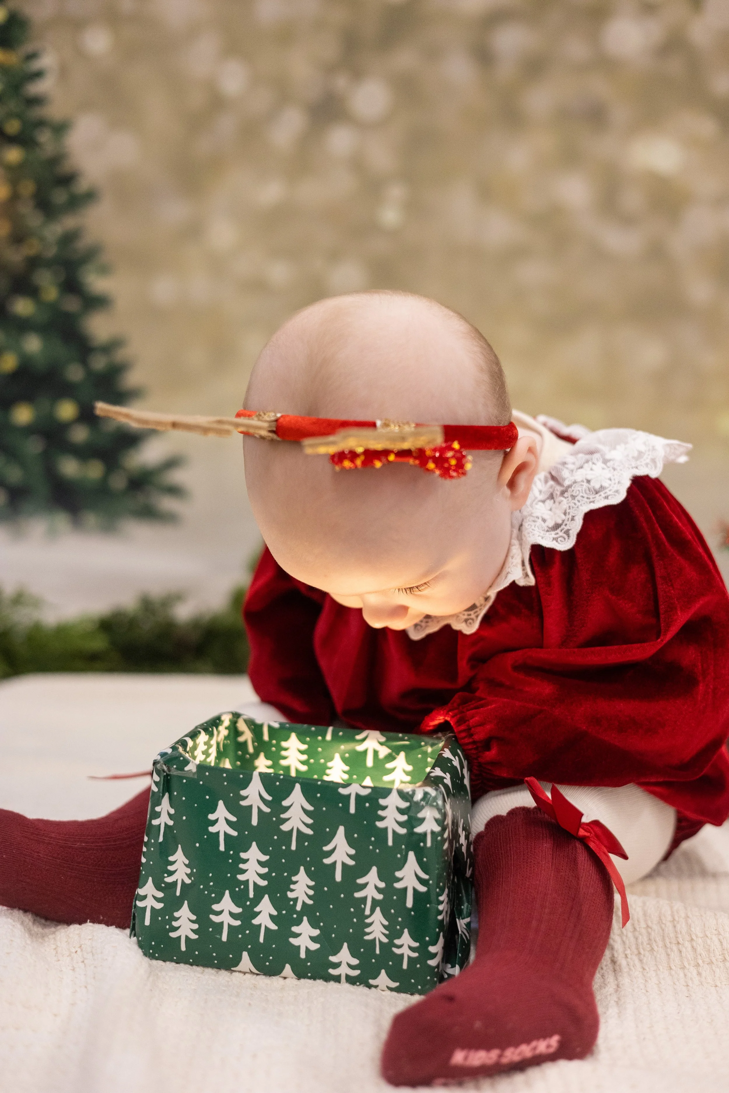 A baby dressed in a red velvet dress and burgundy stockings with a bow, leaning forward and reaching into a Christmas gift bag with a pattern of white Christmas trees. In the background, a blurred Christmas tree with ornaments is visible.