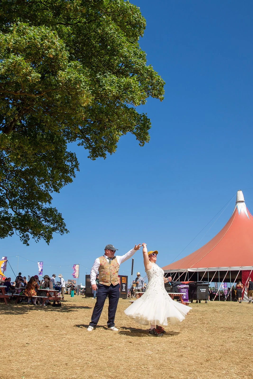 A man and woman dancing outdoors at a festival near a large orange tent, with festivalgoers sitting at picnic tables and a large green tree in the background, under a bright blue sky.