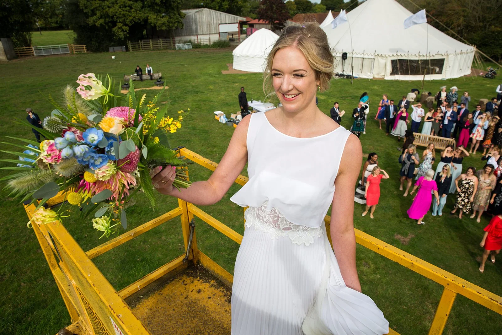 A woman in a white dress holding a colorful bouquet of flowers at an outdoor event or celebration, with people gathered in the background near a large white tent.