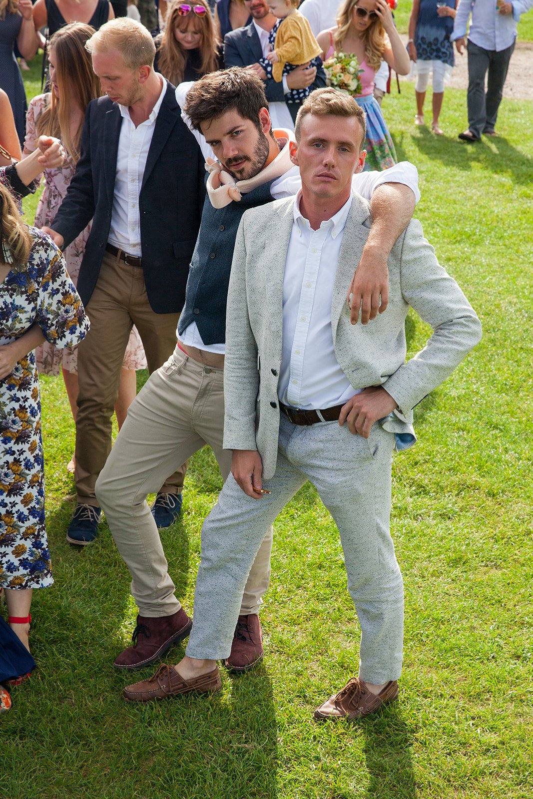 A group of people gathered outdoors in a grassy area, with two young men in the foreground striking confident poses. One man is dressed in a light gray suit, and the other in casual attire with a vest and hoodie. The background shows more people, som