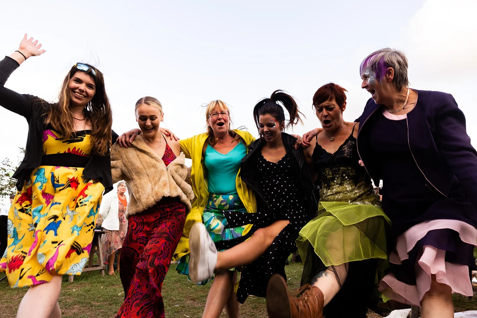 Group of women standing in a circle outdoors, smiling and holding each other, dressed in colorful, patterned clothing, appear to be celebrating or participating in a lively event.