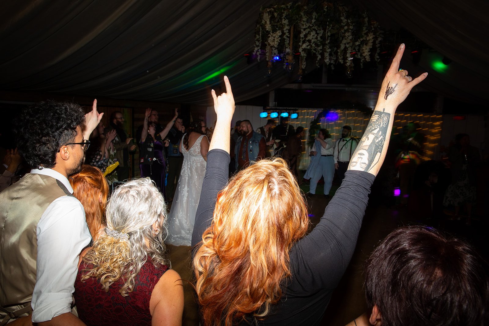 People dancing at a lively indoor celebration, with some raising their hands in the air, under a decorated ceiling with hanging flowers.