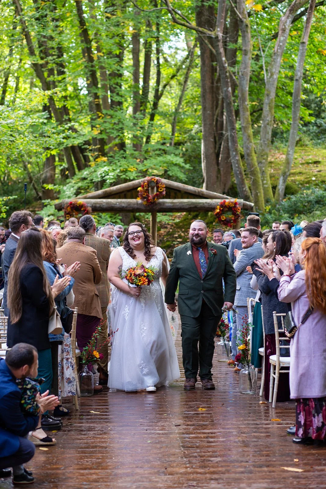 A couple is walking down the aisle during a wedding ceremony outdoors in a forest, surrounded by smiling guests clapping, with a wooden arch decorated with flowers in the background.