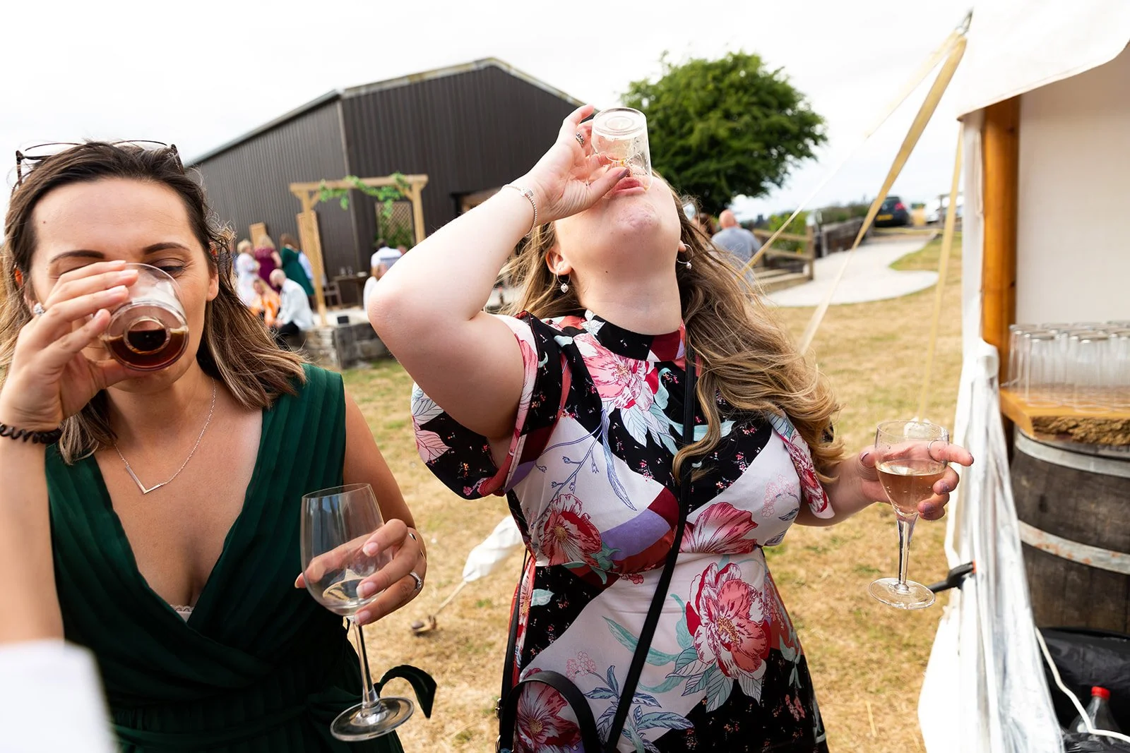 Two women in formal dresses drinking from glasses at an outdoor event with a barn and people in the background.