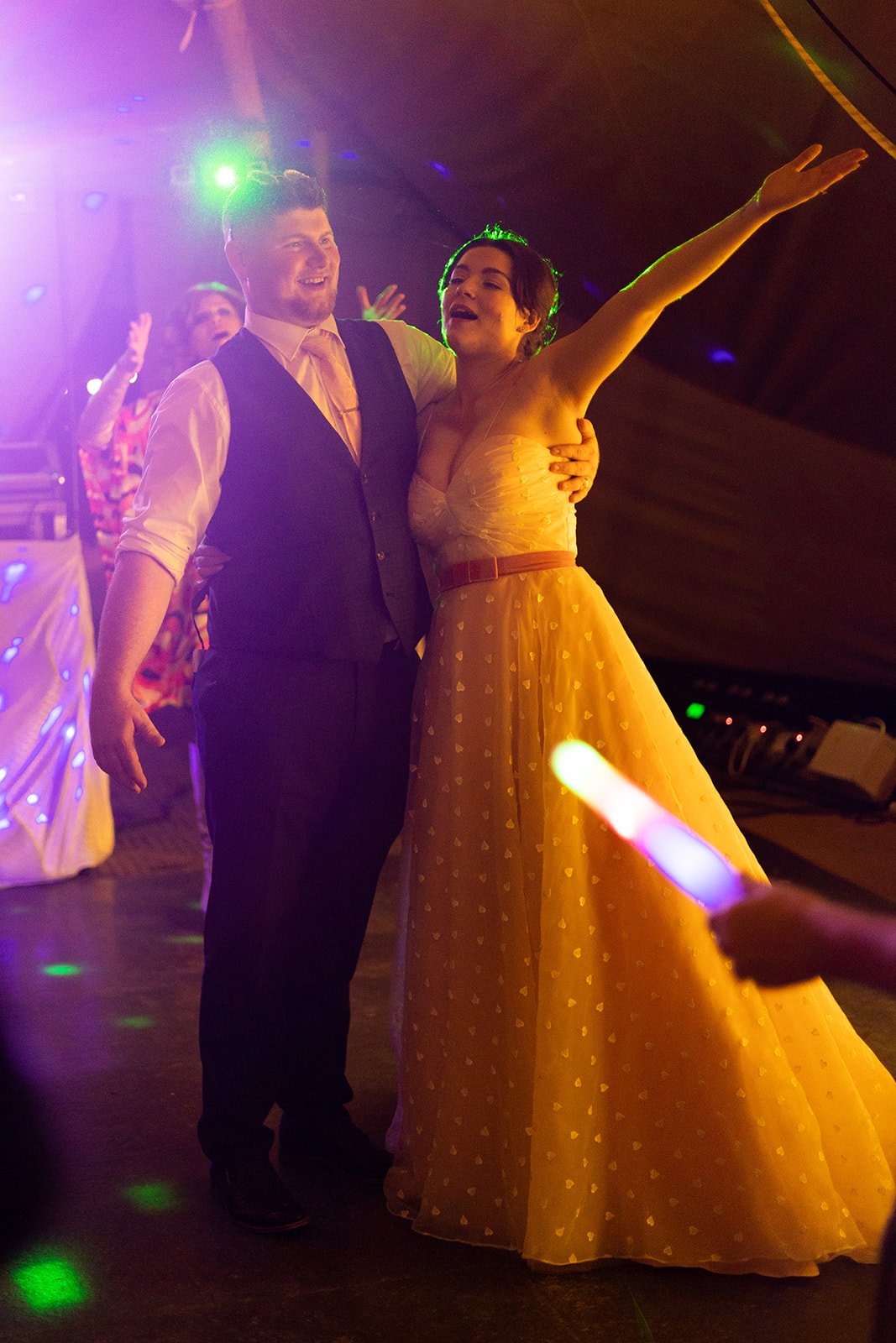 People dancing at a party, with two individuals in the foreground smiling and raising their arms, colorful lighting, and a person holding a glow stick.