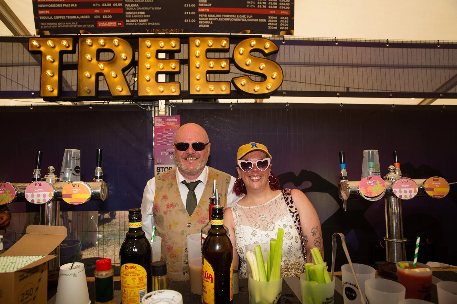 Two people standing behind a bar with beer taps, smiling for the camera. The man is wearing sunglasses, a white shirt, and a floral vest; the woman is wearing heart-shaped sunglasses, a yellow cap, and a patterned top. The bar has various bottles, co