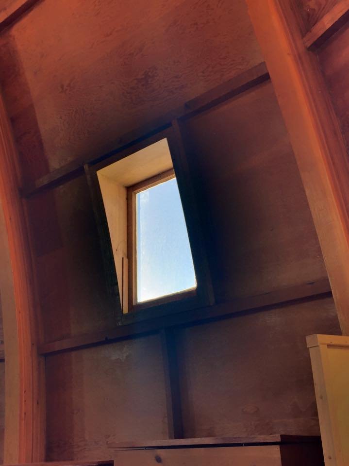  One section of the wall of a tall wooden dome seen from the inside, with a vertical rectangular porthole letting in daylight. 