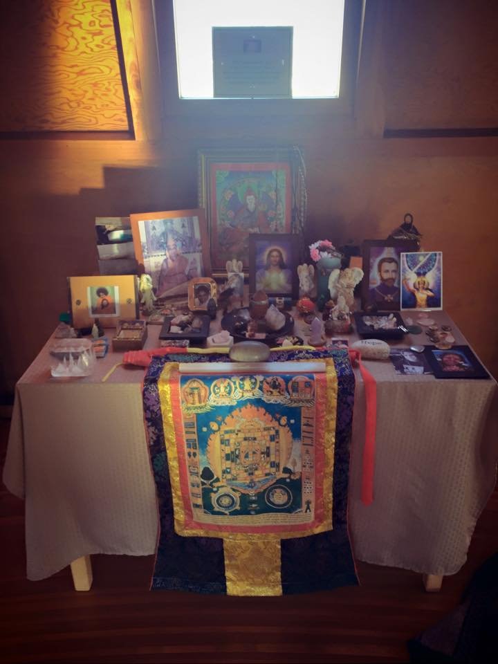  A crowded altar table with a Buddhist thangka cloth draped in front, with photos, icons, and objects from a variety of traditions placed on it. 