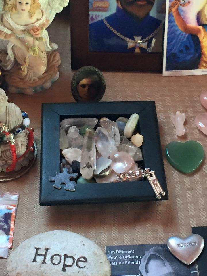  Close-up of a box of gemstones on an altar table, with a jigsaw puzzle piece on one corner of the rim and a sterling silver Torah scroll pendant and chain on another corner. At the bottom, a round rock with “Hope” carved into it and a metallic heart