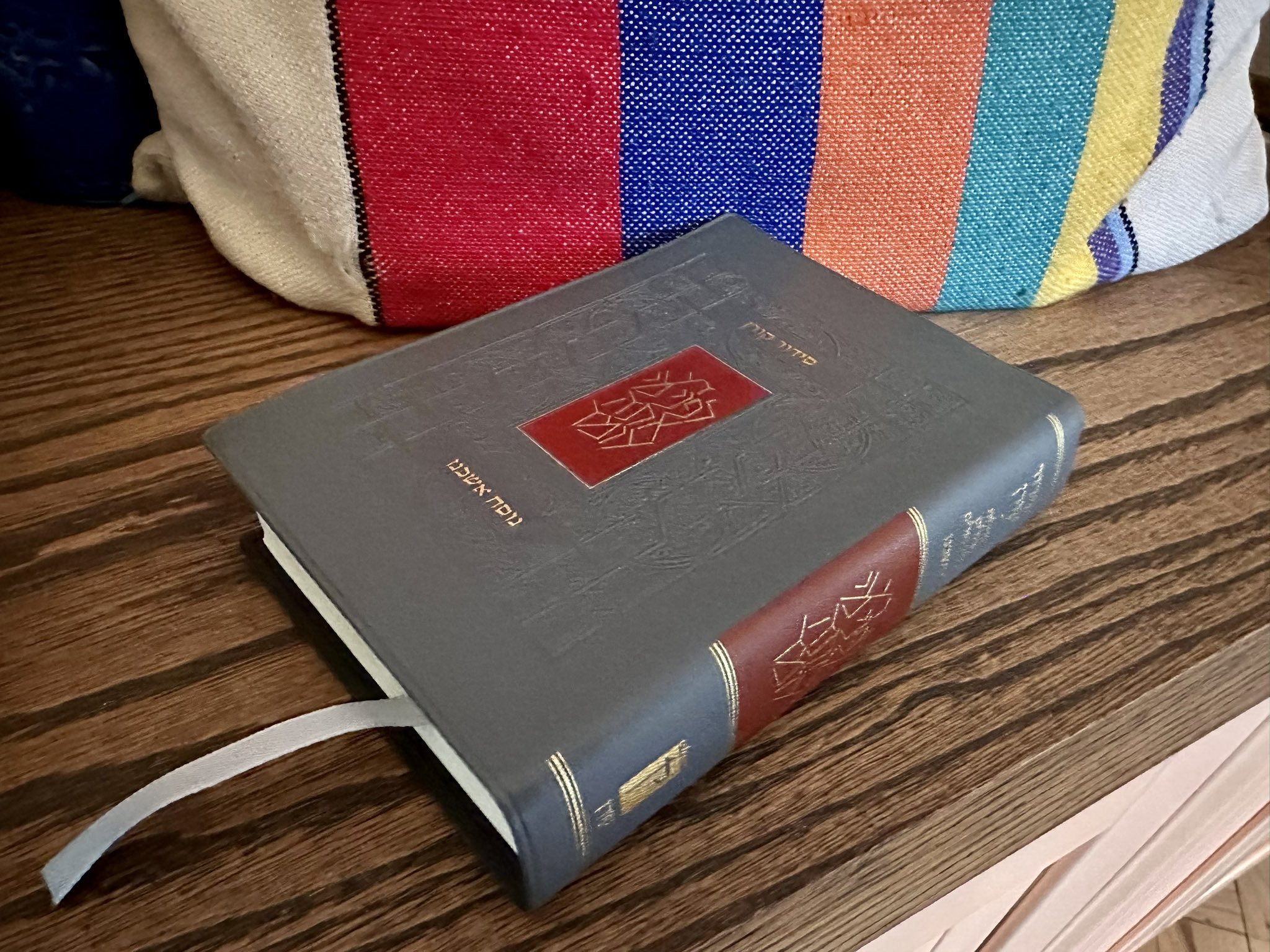 A gray siddur on a wooden counter in front of a colorful tallit bag.