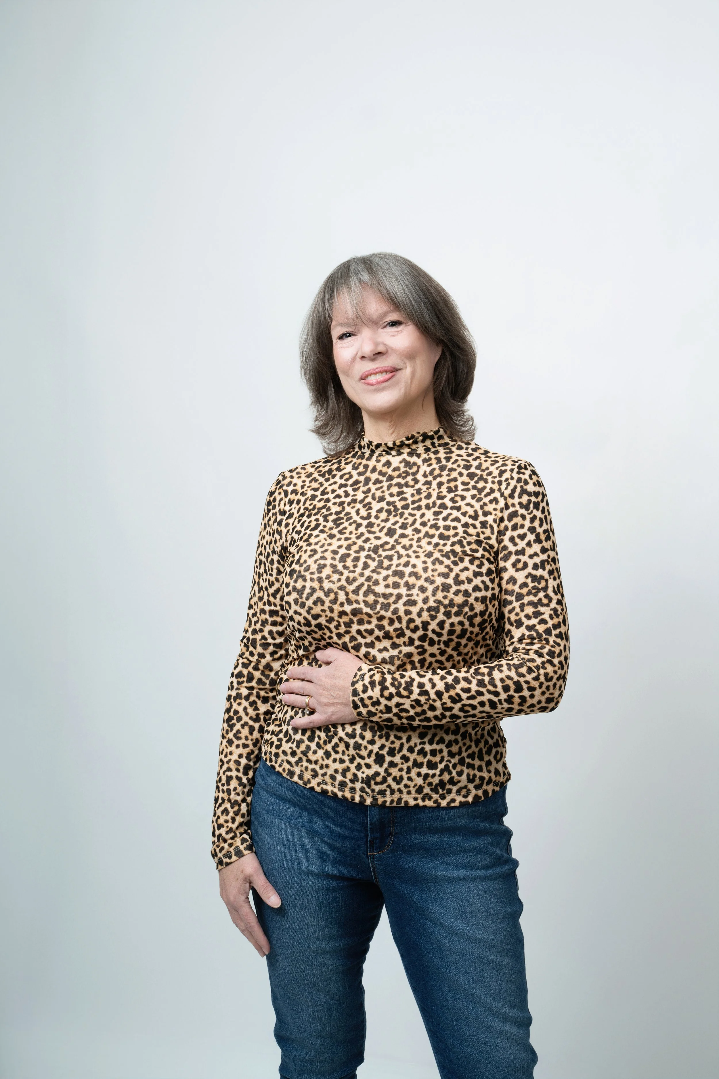 A woman wearing a leopard-print long-sleeve shirt and jeans standing against a plain white background.