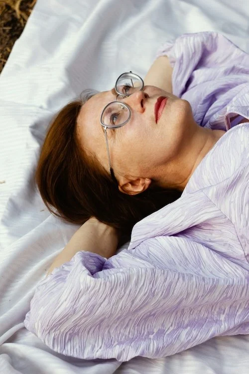 A woman with glasses lying on a bed, looking up.