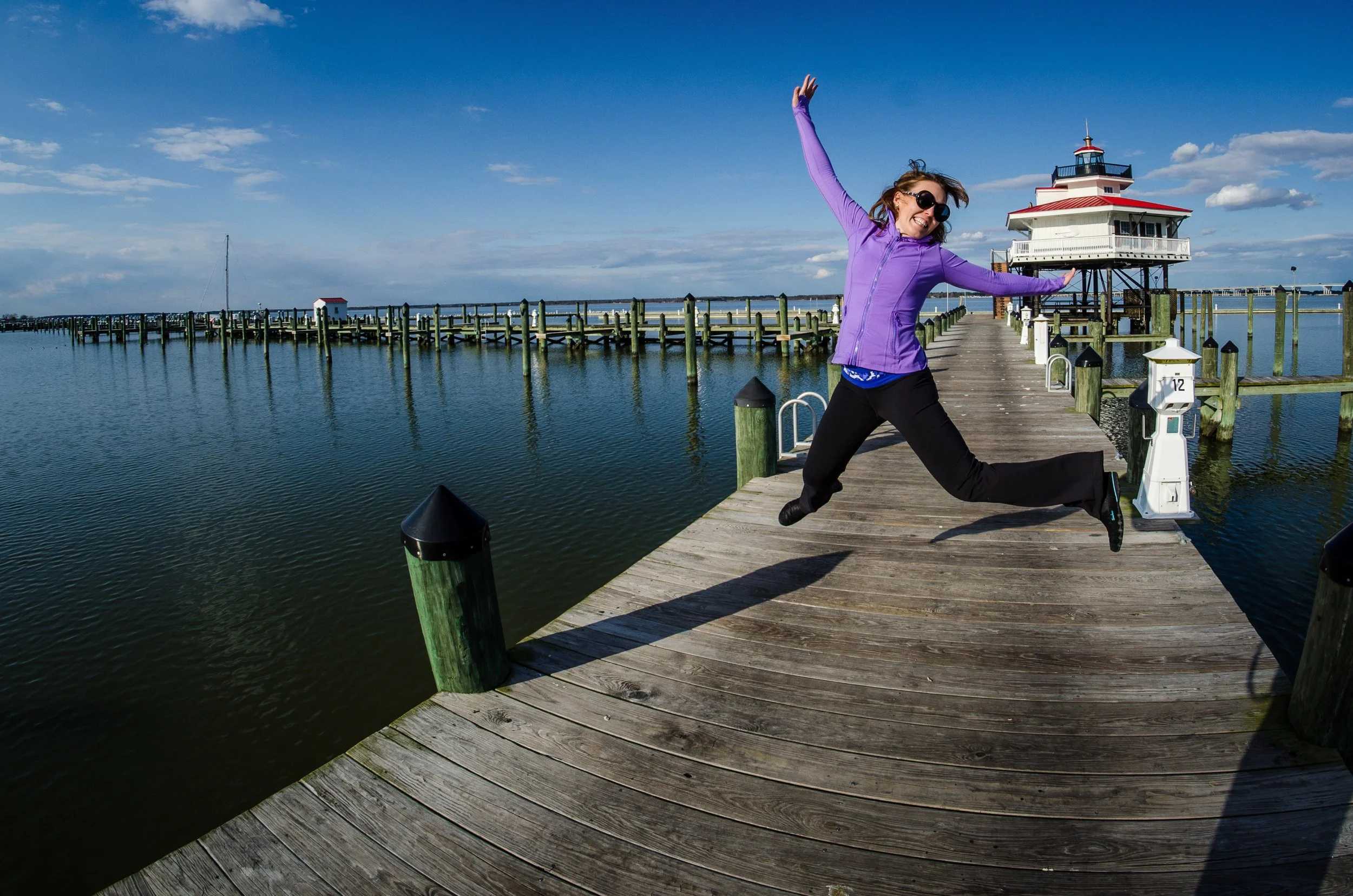Woman in purple jacket jumping on a wooden dock over calm water with a lighthouse in the background.