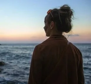 A woman standing by the ocean at sunset, facing away from the camera with her hair in a bun and sunglasses on her head.