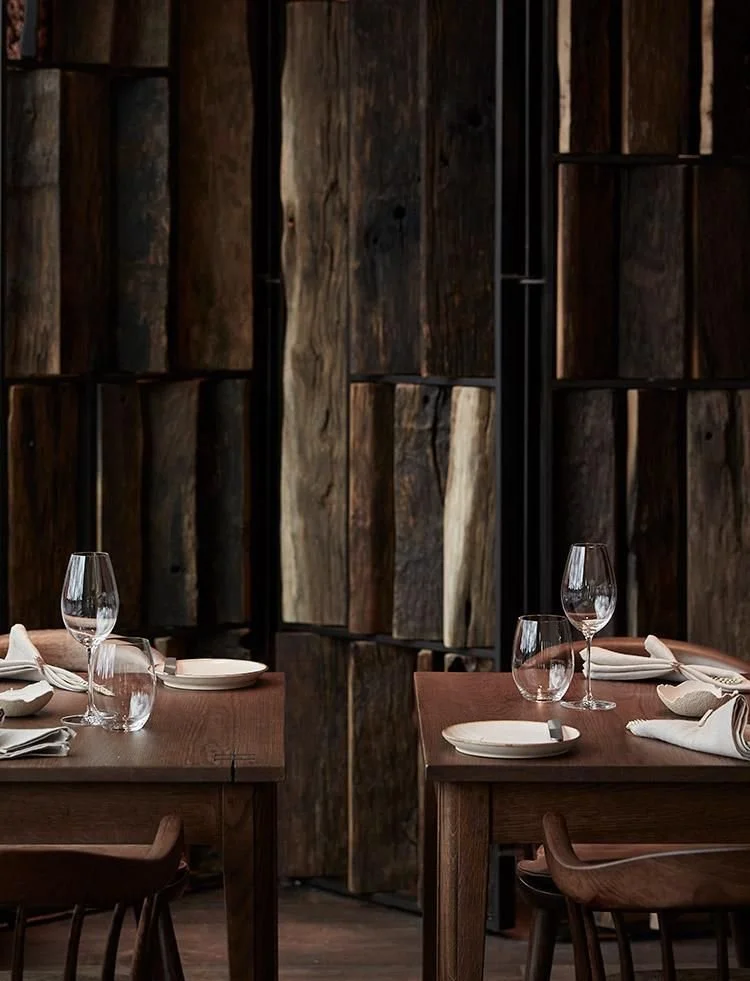 Restaurant with wooden tables set for a meal, including wine glasses, plates, and napkins, against a textured wooden wall.
