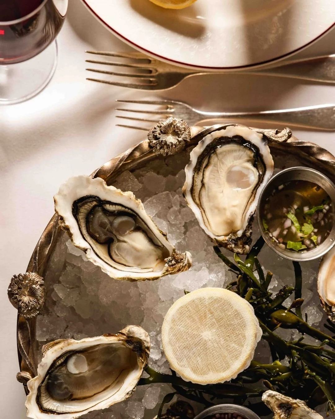 A seafood platter with fresh oysters on ice, garnished with seaweed and accompanied by lemon slices and a small container of mignonette sauce, served on a silver tray.