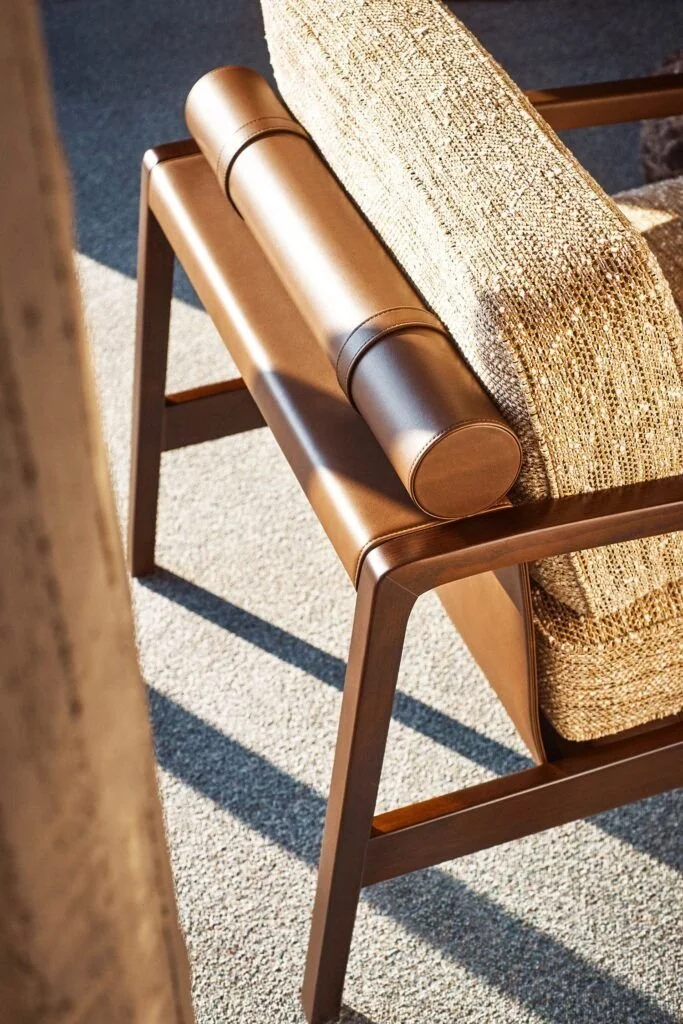 Close-up of a wooden chair with a cylindrical brown bolster pillow resting on its armrest, next to a textured beige cushion.