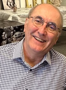A smiling man with glasses and a checkered shirt, sitting indoors with a decorative background.