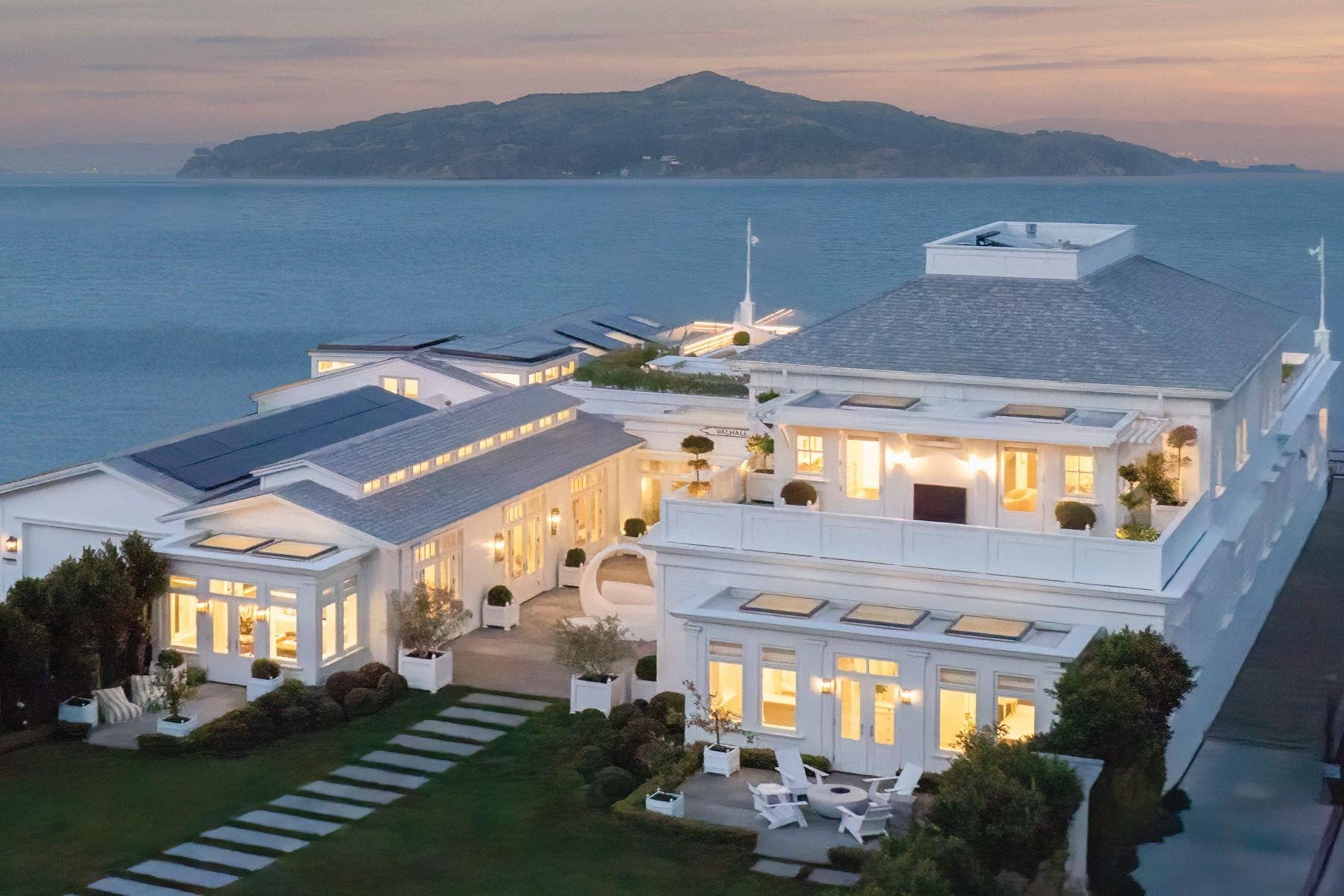 An aerial view of a luxurious white house by the water at sunset, with illuminated windows, outdoor seating areas, and a hillside in the background.