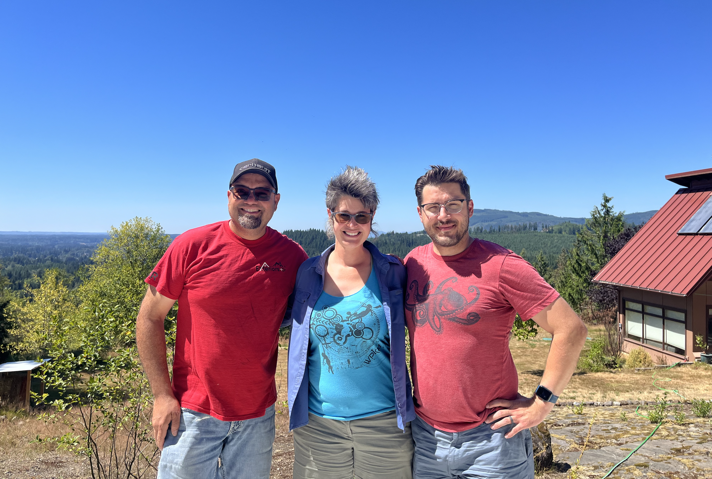 Three people standing outdoors with trees and hills in the background, under a clear blue sky.