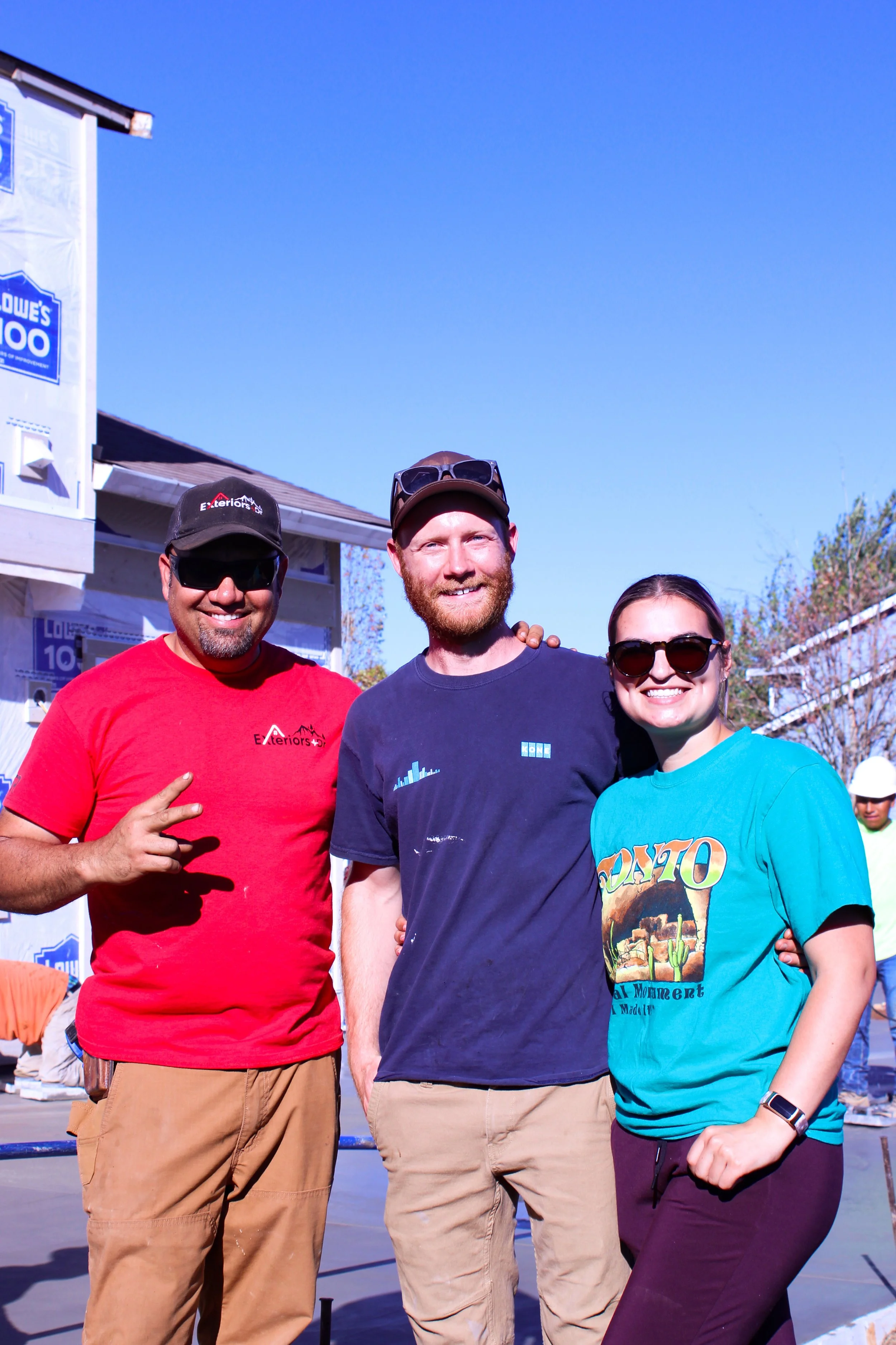 Three people posing outdoors in casual attire, with construction material and a house being built in the background.