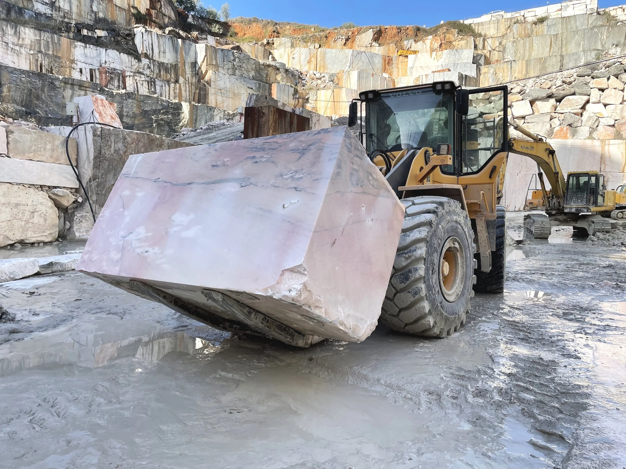 A yellow front loader with a large marble block attached to its bucket at a quarry with stone walls and other construction equipment in the background. Pink marble block