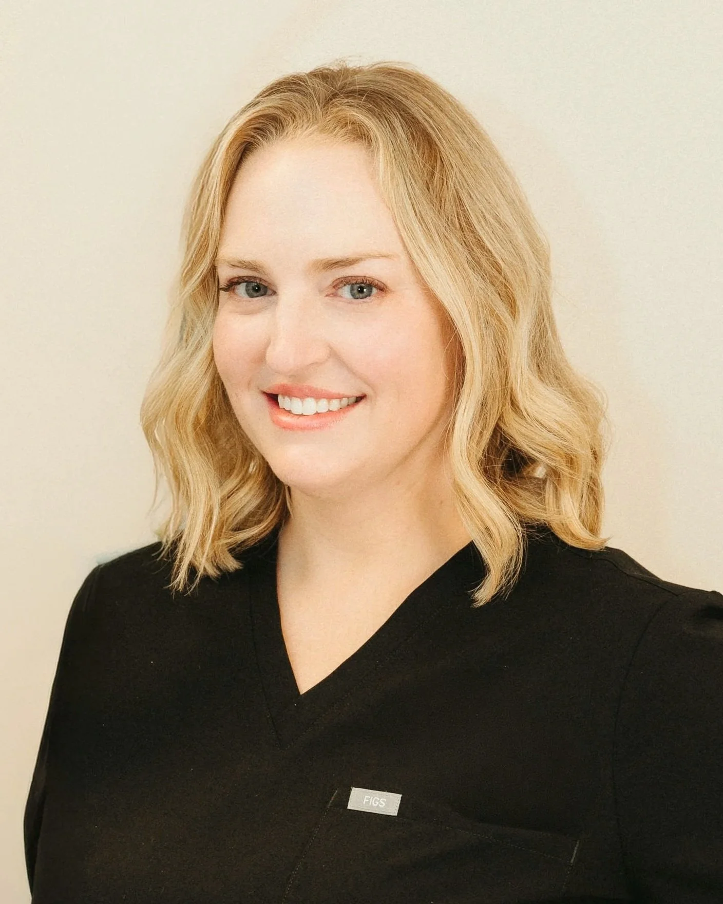 A woman with medium-length blonde hair wearing a black top with a small pin that says 'FIGS' on it, standing against a plain beige background.