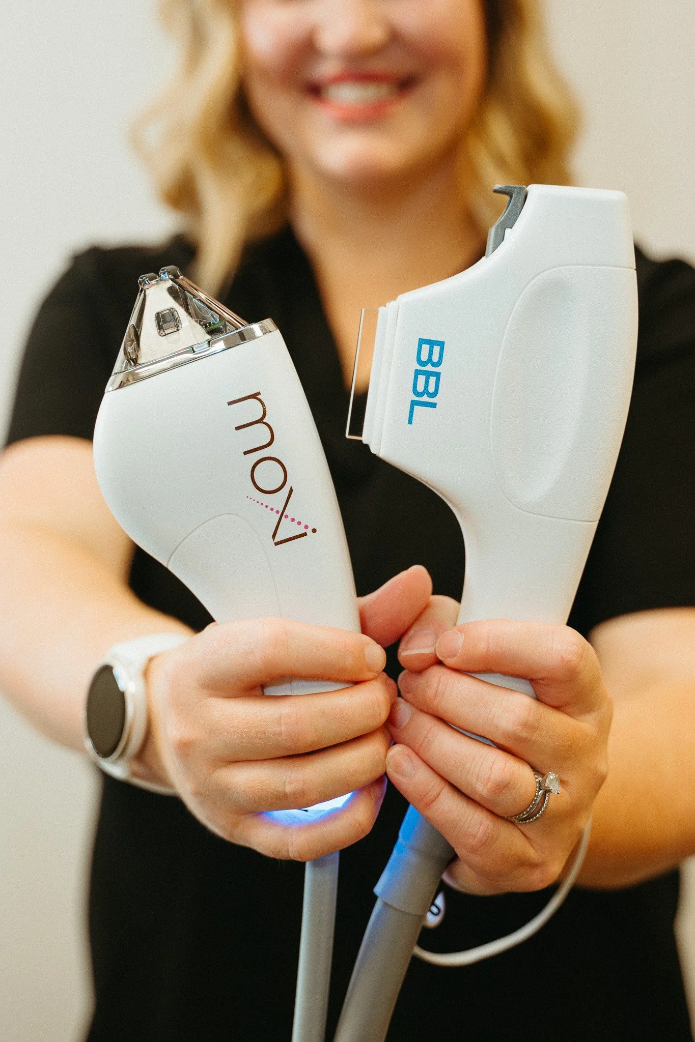 A woman holding two laser hair removal devices, one labeled 'Moxi' and the other labeled 'BBL,' while smiling.