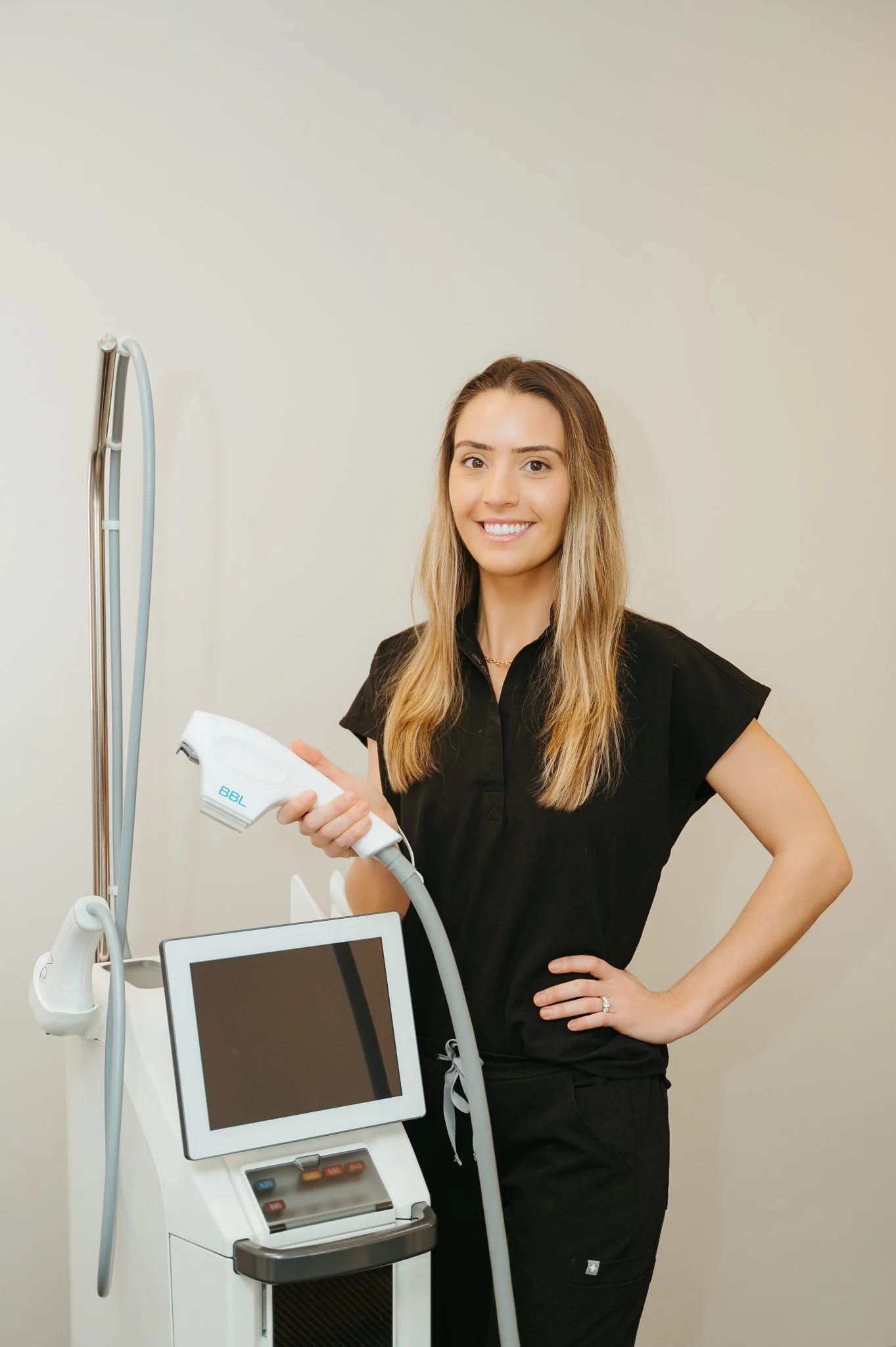 Nurse Olivia Stucker - A smiling woman with long blonde hair, wearing a black scrub top, standing next to medical laser equipment, holding a handheld device, in a clinical setting.