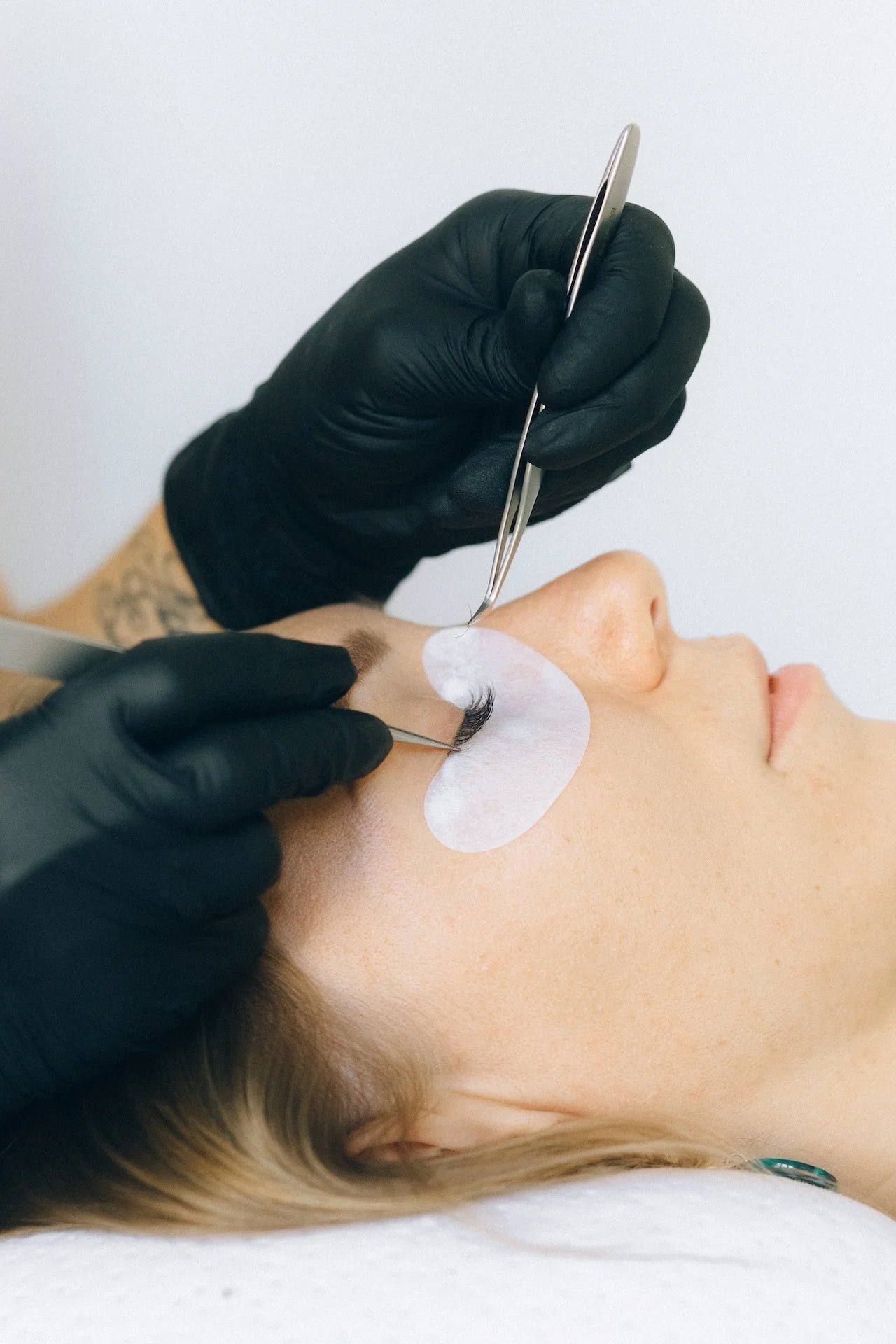 A person receiving eyelash extension application on their right eye by an esthetician wearing black gloves.